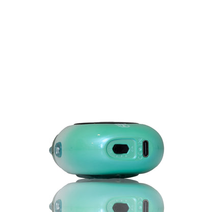 A close-up of the side of a glossy, rounded electronic device in a mint green color, showing a port and a small circular detail, with a reflection visible below on a white background.